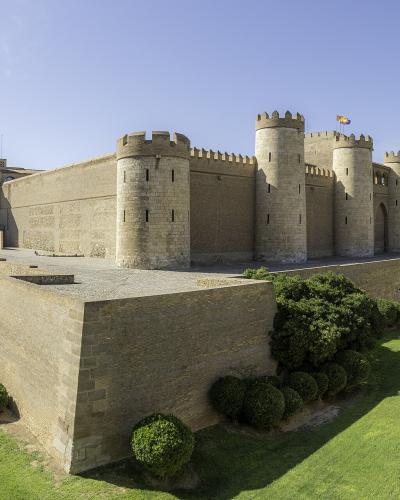 A large medieval fortress with crenellated stone walls, round towers, and a flag flying from the highest tower, surrounded by manicured green lawns and hedges under a clear blue sky.
