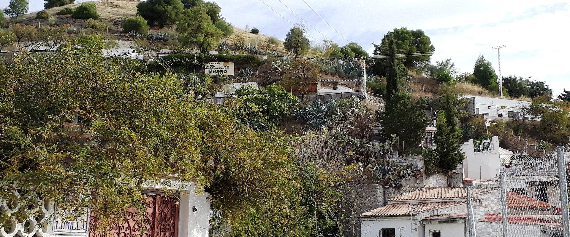 The image shows a hillside scene with white-walled buildings, terracotta roofs, and lush vegetation, including a prominent gate on the left and a person standing on a stone wall in the foreground.