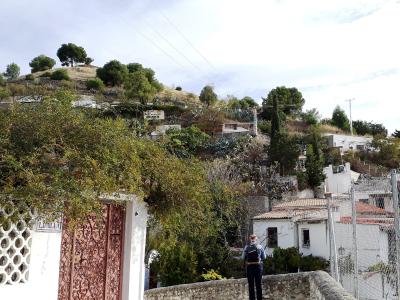 The image shows a hillside scene with white-walled buildings, terracotta roofs, and lush vegetation, including a prominent gate on the left and a person standing on a stone wall in the foreground.