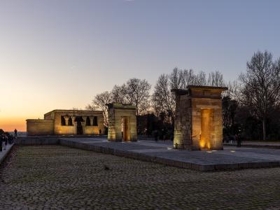 A low-angle evening photograph of an ancient Egyptian-style stone temple complex with a pylon gate and main sanctuary building, illuminated by artificial lights against a twilight sky, situated in a park with cobblestone paths and bare trees.