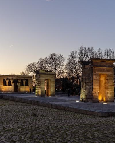 A low-angle evening photograph of an ancient Egyptian-style stone temple complex with a pylon gate and main sanctuary building, illuminated by artificial lights against a twilight sky, situated in a park with cobblestone paths and bare trees.