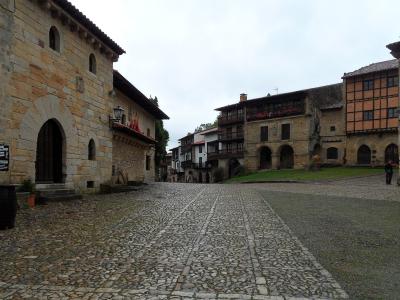 A cobblestone town square surrounded by historic stone and half-timbered buildings, including one with a large arched entrance and another with wooden balconies; a person walks on the right side under an overcast sky.