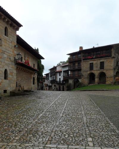 A cobblestone town square surrounded by historic stone and half-timbered buildings, including one with a large arched entrance and another with wooden balconies; a person walks on the right side under an overcast sky.
