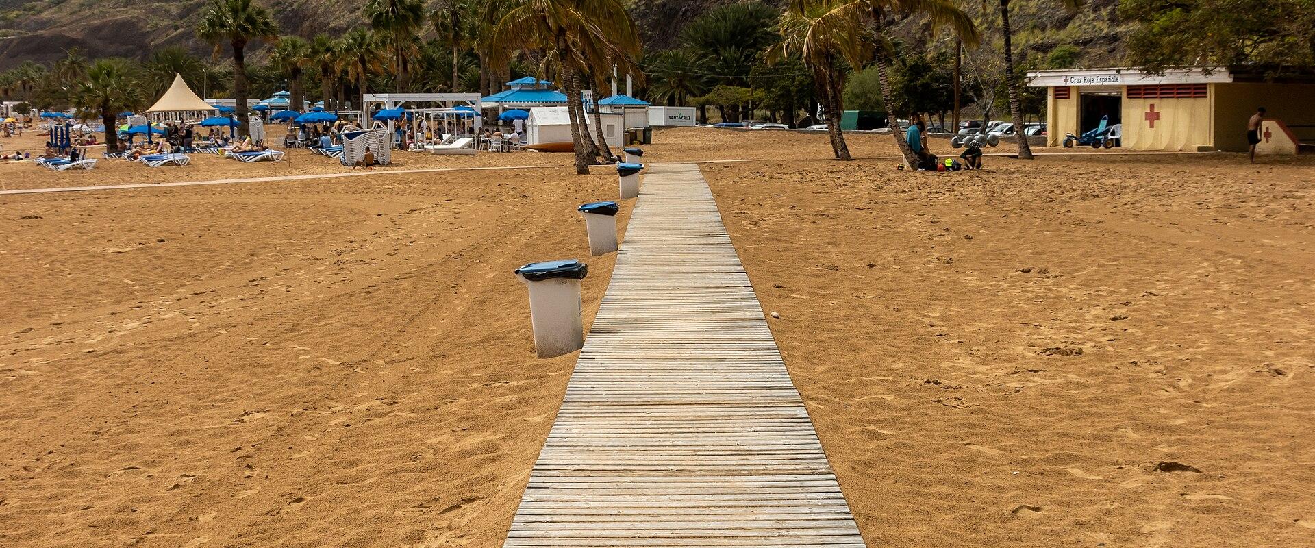 A wooden boardwalk extends across golden sand towards a beach area with palm trees, blue umbrellas, and a lifeguard station, backed by steep, rugged mountains under a cloudy sky.