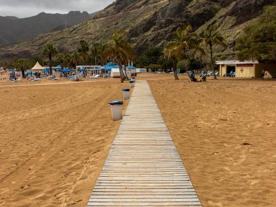 A wooden boardwalk extends across golden sand towards a beach area with palm trees, blue umbrellas, and a lifeguard station, backed by steep, rugged mountains under a cloudy sky.