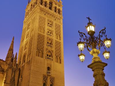 A low-angle shot of the Giralda bell tower of Seville Cathedral at twilight, featuring its distinctive Moorish brickwork, Gothic upper levels, and the La Giraldilla weather vane on top, with an ornate street lamp in the foreground.