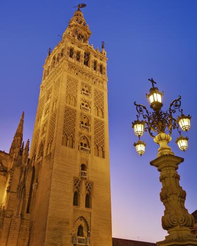 A low-angle shot of the Giralda bell tower of Seville Cathedral at twilight, featuring its distinctive Moorish brickwork, Gothic upper levels, and the La Giraldilla weather vane on top, with an ornate street lamp in the foreground.
