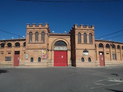 A brick bullring facade with two crenellated towers, a central arched entrance with red doors, and a sign reading "PLAZA DE TOROS" above the entrance, set against a clear blue sky.