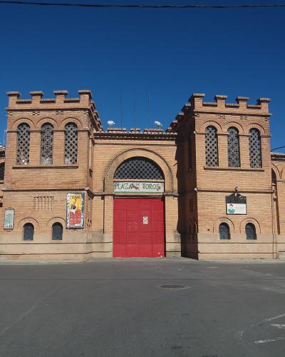 A brick bullring facade with two crenellated towers, a central arched entrance with red doors, and a sign reading "PLAZA DE TOROS" above the entrance, set against a clear blue sky.