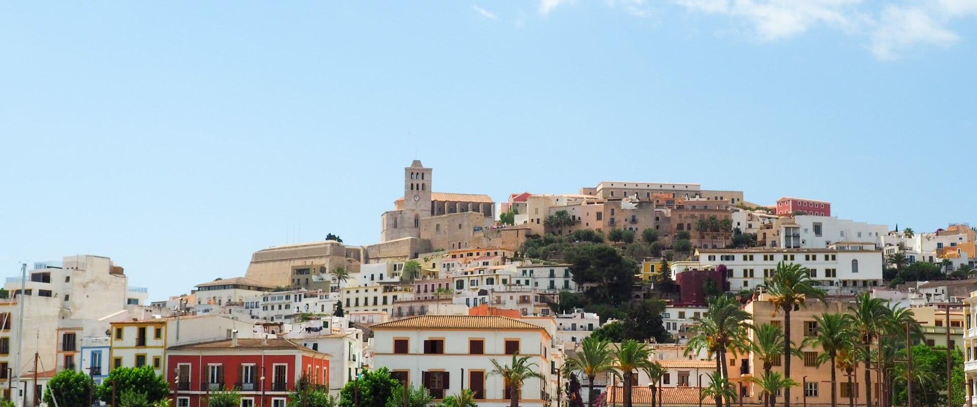 A view of the historic walled town of Ibiza (Dalt Vila) rising on a hill above a harbor promenade lined with palm trees and boats.