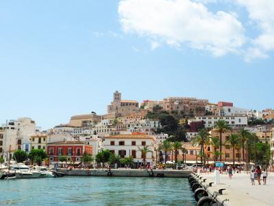 A view of the historic walled town of Ibiza (Dalt Vila) rising on a hill above a harbor promenade lined with palm trees and boats.