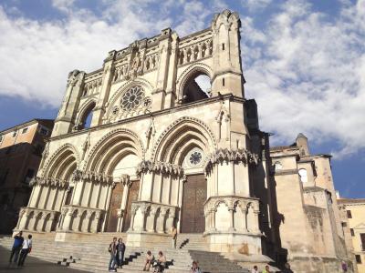 A low-angle photograph of a large Gothic cathedral facade with three prominent arched portals, a rose window, and a distinctive open upper gallery, with people sitting on the steps in the foreground.