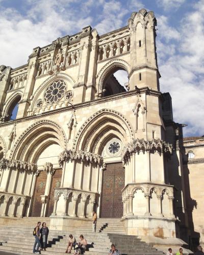 A low-angle photograph of a large Gothic cathedral facade with three prominent arched portals, a rose window, and a distinctive open upper gallery, with people sitting on the steps in the foreground.