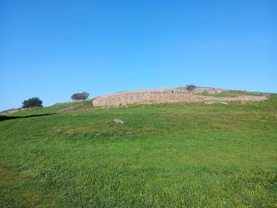 A grassy hillside with a low, curved stone retaining wall or ruin running along the ridge under a clear blue sky.