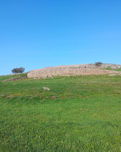 A grassy hillside with a low, curved stone retaining wall or ruin running along the ridge under a clear blue sky.