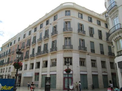 A low-angle view of a large, multi-story cream-colored building with a distinctive curved corner facade, featuring numerous windows with dark shutters and wrought-iron balconies, situated on a pedestrian street with lampposts and people.