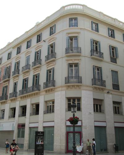 A low-angle view of a large, multi-story cream-colored building with a distinctive curved corner facade, featuring numerous windows with dark shutters and wrought-iron balconies, situated on a pedestrian street with lampposts and people.