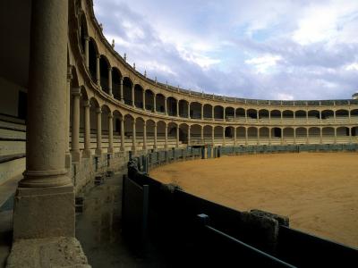 The image shows the interior of a historic bullring with a curved facade of stone arches and columns overlooking a sandy arena floor.