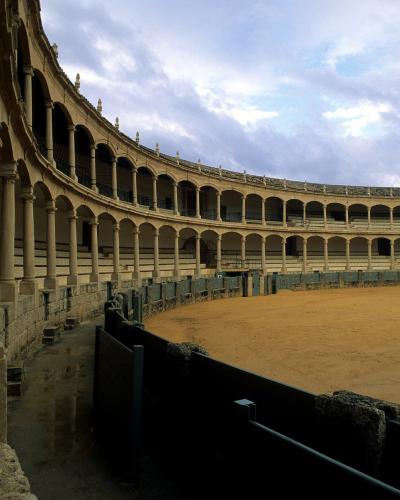 The image shows the interior of a historic bullring with a curved facade of stone arches and columns overlooking a sandy arena floor.