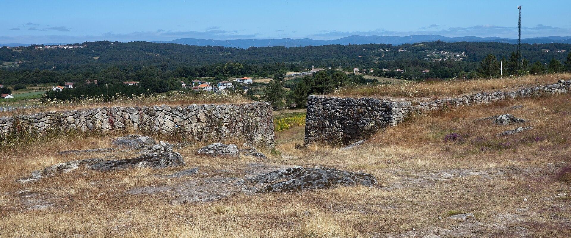 A view from an ancient stone-walled archaeological site (a Castro) overlooking a green, hilly valley with scattered houses and a distant radio tower under a blue sky.
