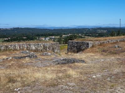 A view from an ancient stone-walled archaeological site (a Castro) overlooking a green, hilly valley with scattered houses and a distant radio tower under a blue sky.