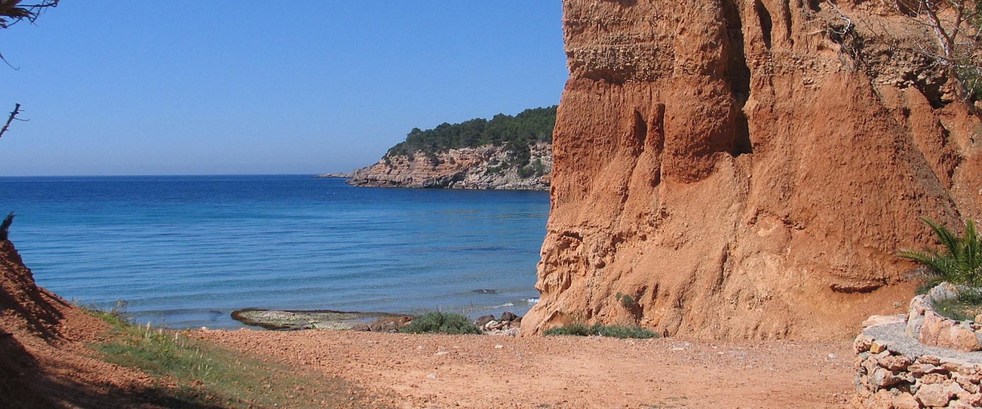 A coastal scene featuring a reddish-orange sandy beach, a steep eroded cliff face on the right, and the blue Mediterranean Sea under a clear sky.