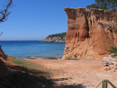 A coastal scene featuring a reddish-orange sandy beach, a steep eroded cliff face on the right, and the blue Mediterranean Sea under a clear sky.