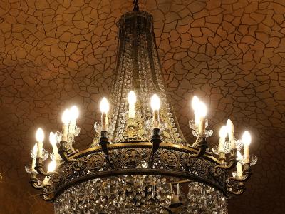 An ornate crystal chandelier hangs from a ceiling with a distinctive mosaic tile pattern, above a stained-glass window with organic, bone-like framing.