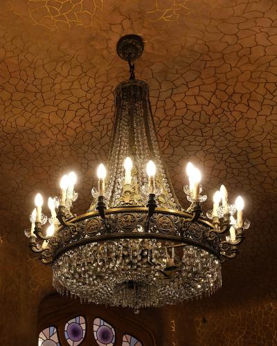An ornate crystal chandelier hangs from a ceiling with a distinctive mosaic tile pattern, above a stained-glass window with organic, bone-like framing.