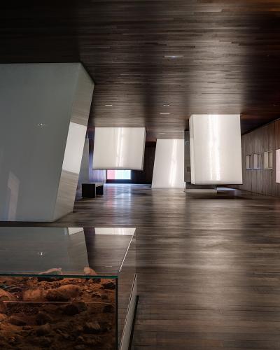 An interior view of a modern museum gallery featuring dark wood flooring and ceiling, large white suspended display panels, and a glass vitrine containing an archaeological excavation in the foreground.