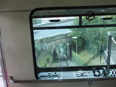 View from inside a funicular car looking out the front window onto the tracks, showing a stone tunnel entrance, overhead road, and control panel.