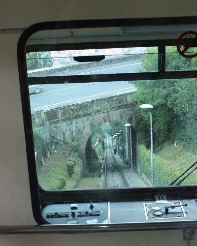 View from inside a funicular car looking out the front window onto the tracks, showing a stone tunnel entrance, overhead road, and control panel.