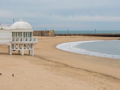A white, domed beach pavilion (chiringuito) on stilts stands on a sandy beach next to a stone breakwater and the ocean under an overcast sky.