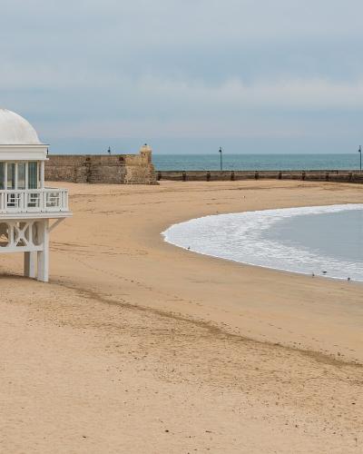 A white, domed beach pavilion (chiringuito) on stilts stands on a sandy beach next to a stone breakwater and the ocean under an overcast sky.