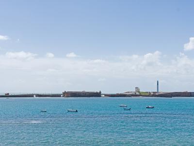 A coastal view of two historic stone fortresses on a peninsula or island connected by a breakwater, with a lighthouse and small boats in the foreground water.