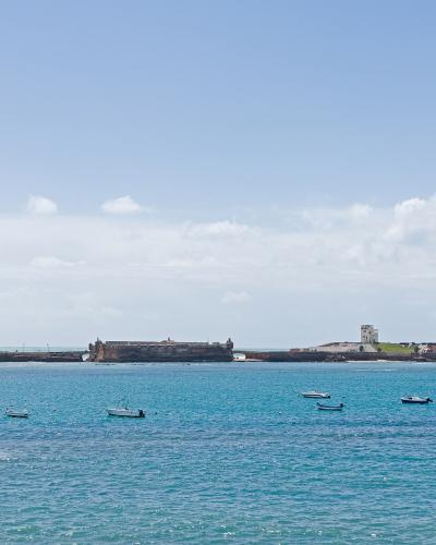 A coastal view of two historic stone fortresses on a peninsula or island connected by a breakwater, with a lighthouse and small boats in the foreground water.