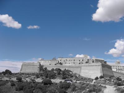 A large stone fortress with high bastions and ramparts sits atop a hill, featuring a prominent church with a dome on the right side, set against a blue sky with selective colorization (sky and clouds in color, landscape in grayscale).