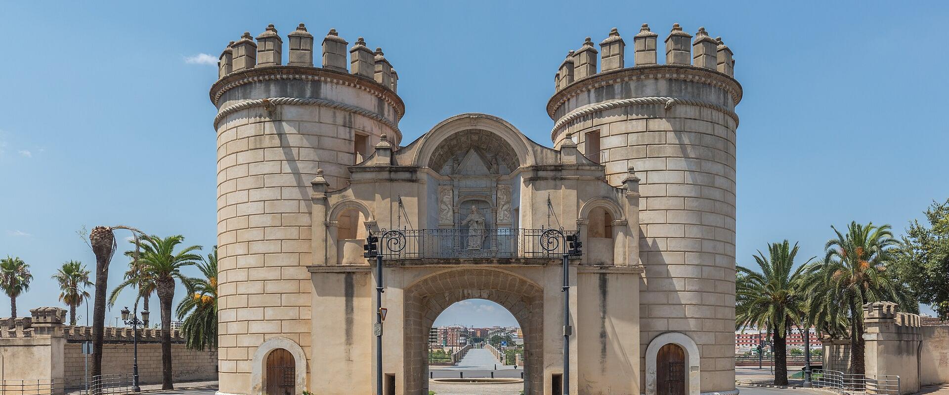 The image shows the Puerta de Palmas, a monumental city gate with two large cylindrical towers and a central archway, located in Badajoz, Spain.
