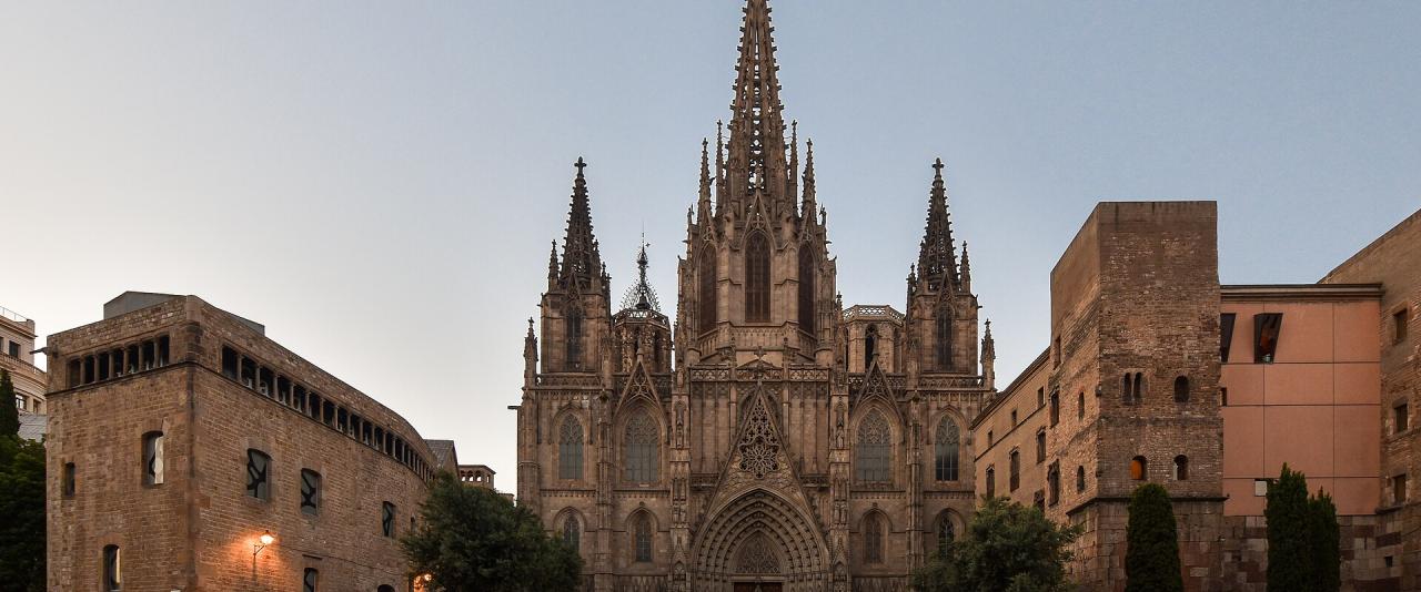 A large Gothic cathedral with a prominent central spire and flanking towers, situated in a paved plaza with surrounding stone buildings and trees.