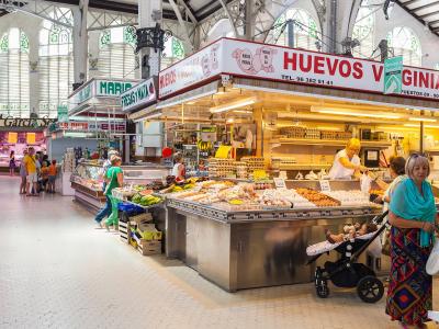 Interior view of a large, historic market hall with high arched windows and metal trusses, featuring stalls selling produce and eggs, with prominent signage reading "HUEVOS VIRGINIA" and "FRESAS Y NATA".