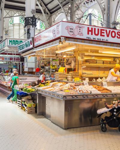 Interior view of a large, historic market hall with high arched windows and metal trusses, featuring stalls selling produce and eggs, with prominent signage reading "HUEVOS VIRGINIA" and "FRESAS Y NATA".