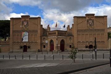 A symmetrical brick building with Mudejar architectural features, including arched gateways and decorative tilework, with a banner on the left tower reading "Escuela de Arte de Toledo".