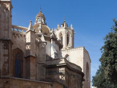 A close-up architectural view of a historic stone cathedral featuring a prominent dome with glazed tiles, Gothic-style windows, and a bell tower against a clear blue sky.