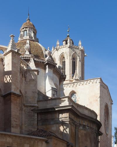 A close-up architectural view of a historic stone cathedral featuring a prominent dome with glazed tiles, Gothic-style windows, and a bell tower against a clear blue sky.