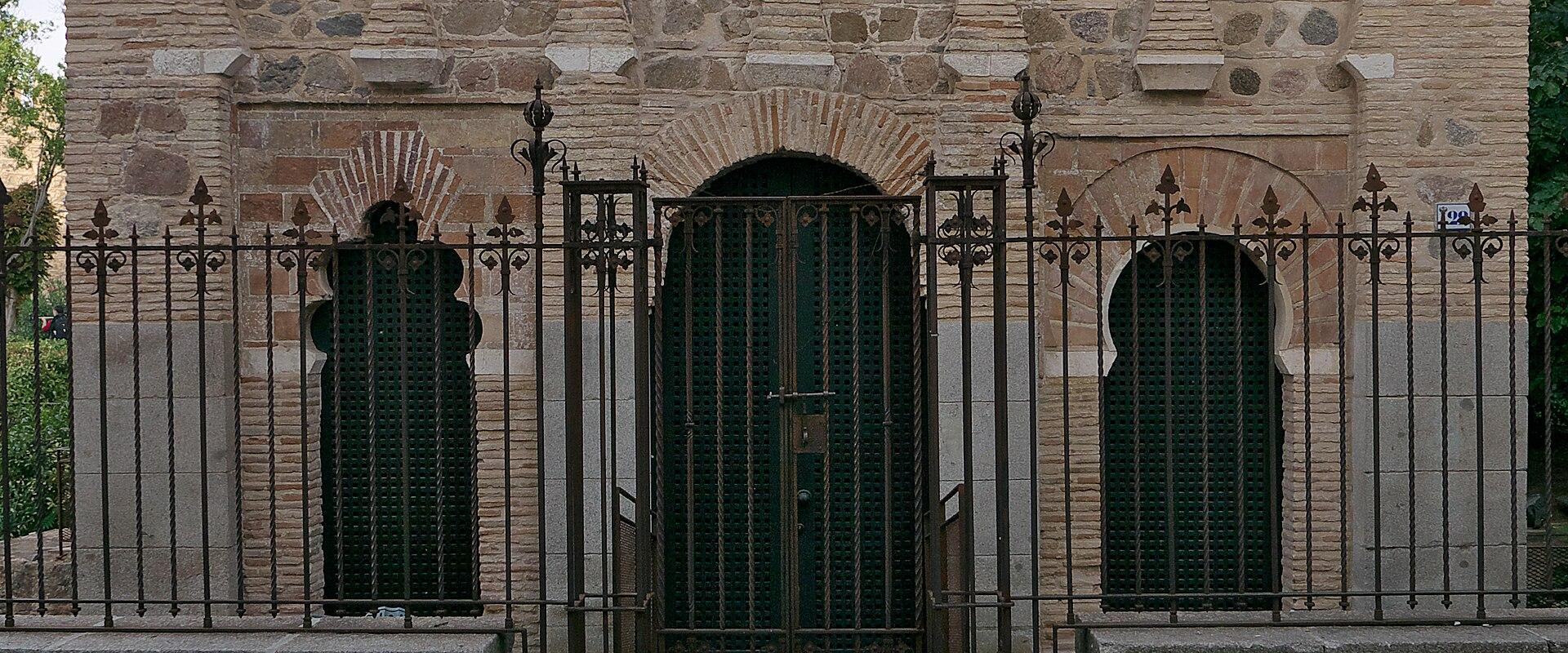 A low-angle photograph of the southwest facade of a small, historic brick building featuring horseshoe arches, a lattice window, and a black metal gate, situated on a cobblestone street.