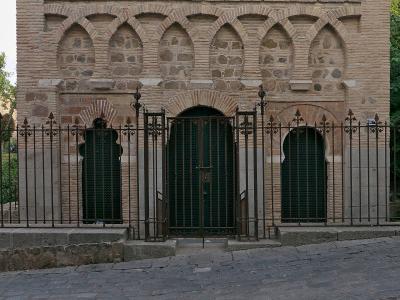A low-angle photograph of the southwest facade of a small, historic brick building featuring horseshoe arches, a lattice window, and a black metal gate, situated on a cobblestone street.