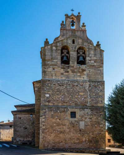 A stone church bell tower with three bells (two in arched openings, one in a smaller upper opening), topped with a cross, set against a clear blue sky with a road and trees nearby.