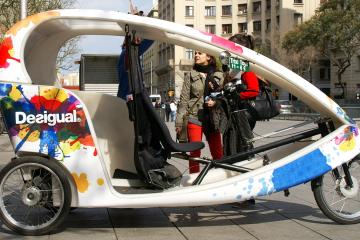 A colorful, three-wheeled pedicab (bicycle taxi) with "Desigual" branding and a "Trixi Tour" sign, parked on a paved city plaza with pedestrians and buildings in the background.