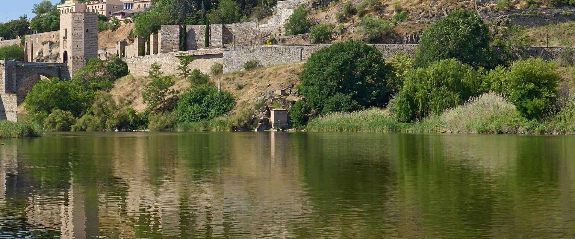 A large, fortified stone palace with distinctive corner turrets sits atop a steep, tree-covered hill overlooking a wide river, with its reflection visible in the water below.