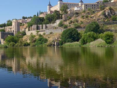 A large, fortified stone palace with distinctive corner turrets sits atop a steep, tree-covered hill overlooking a wide river, with its reflection visible in the water below.